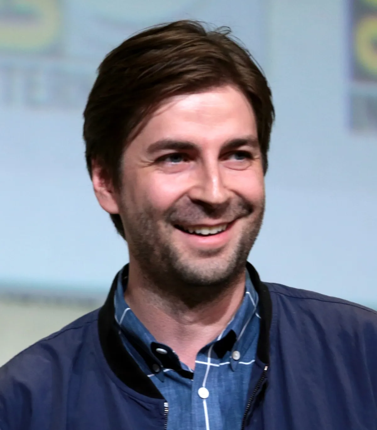 A man with short brown hair and a beard smiles warmly, wearing a dark jacket over a blue, collared shirt, standing against a blurred background with text and graphics.