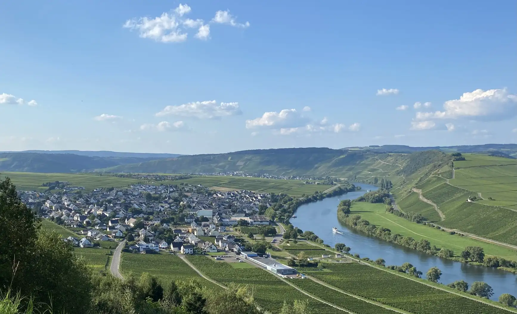 A panoramic view of a river winding through green hills and a small town beneath a bright blue sky.
