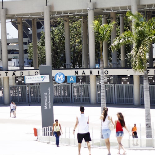 Oamenii merg pe jos în fața Stadionului Maracanã în timpul zilei, cu palmieri și intrarea stadionului vizibile în fundal.