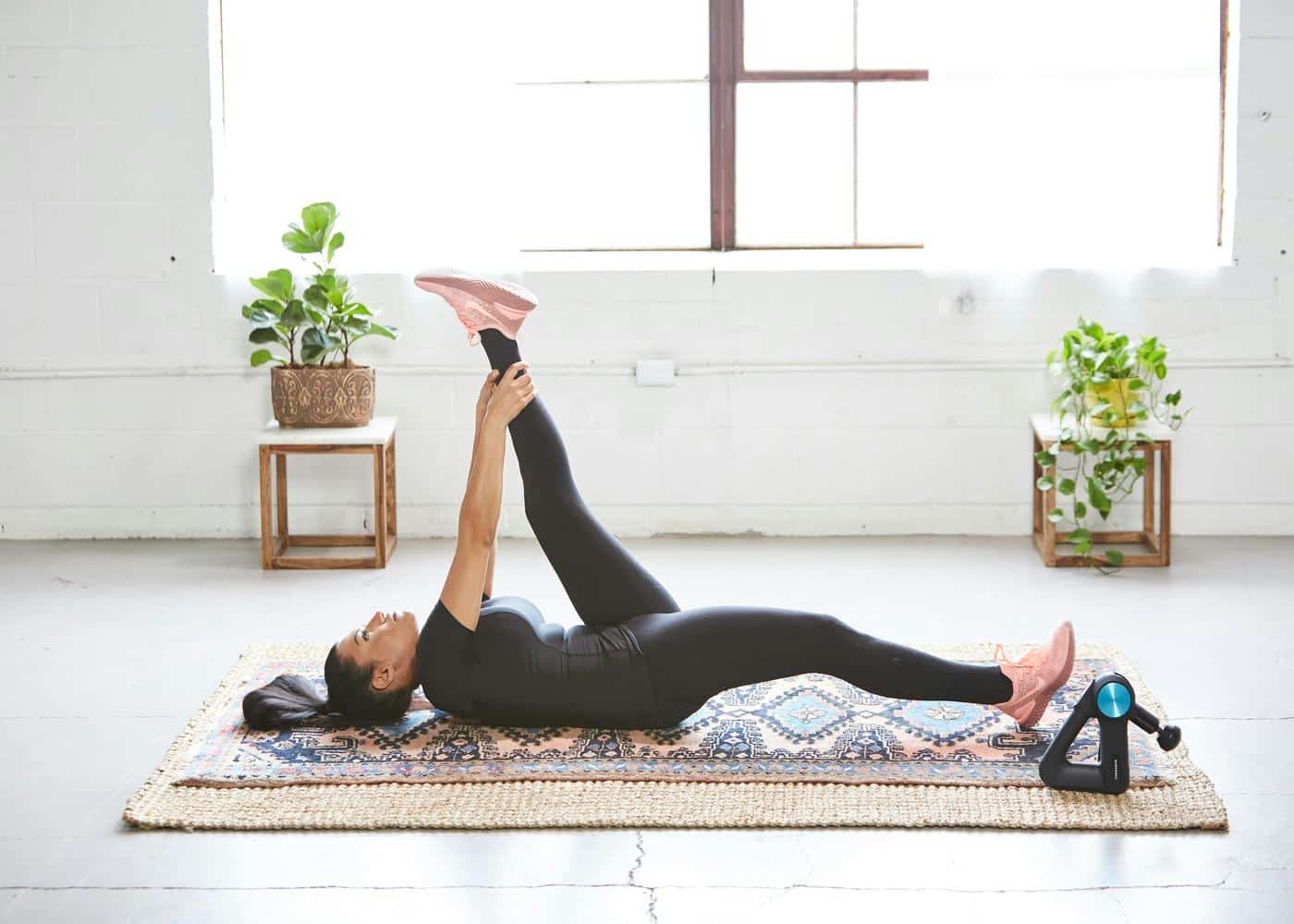 Woman laying on yoga mat stretching her leg in the air with a massage gun on the side