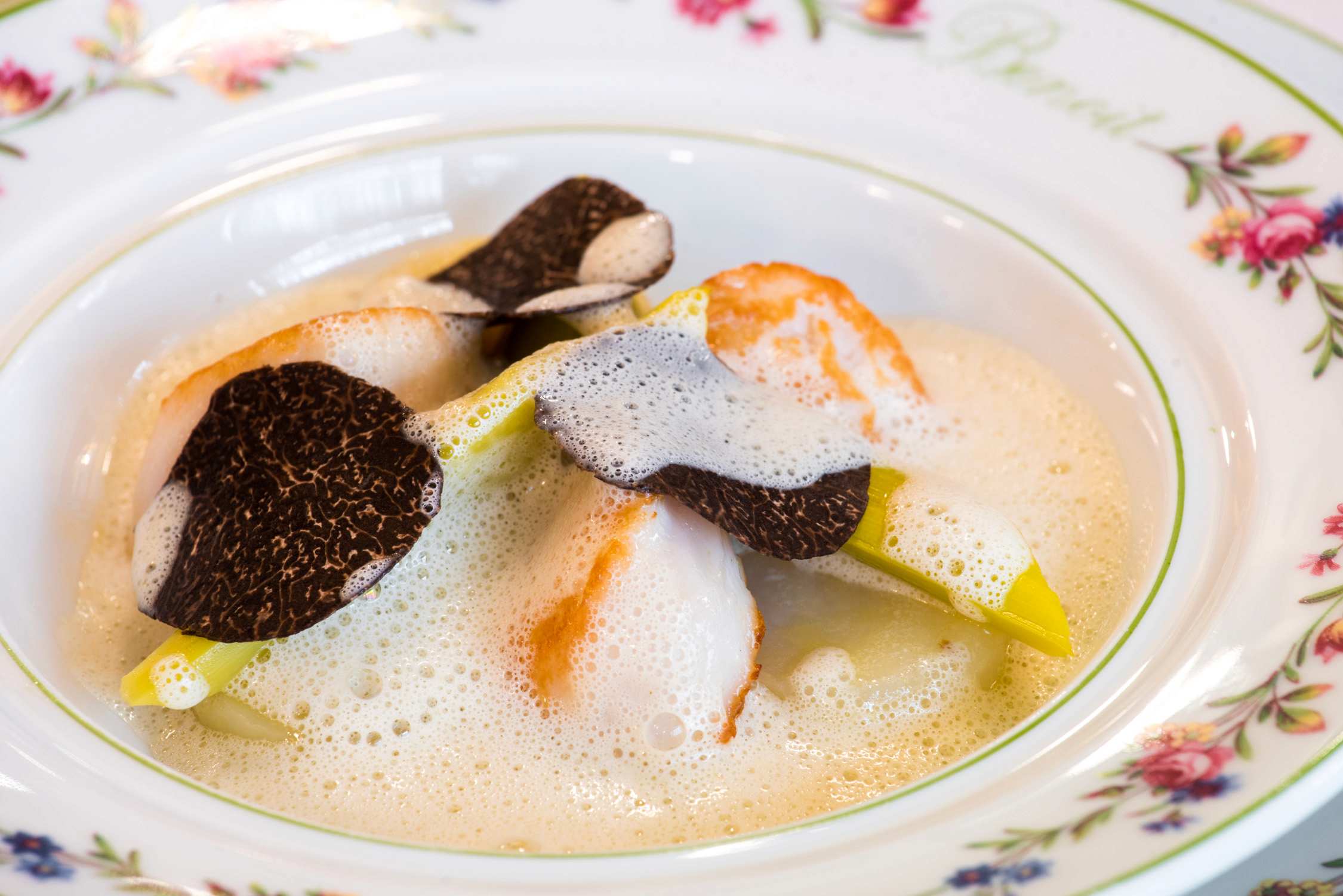 A bowl of creamy soup containing orange segments, yellow fruit slices, and dark taro chips served in a floral dish.