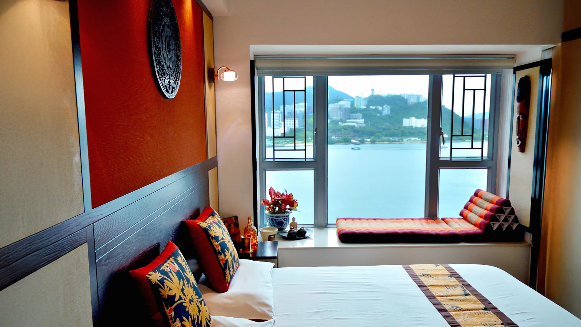 Bedroom with a large window overlooking a waterfront, featuring a dark wood headboard and colourful patterned cushions.