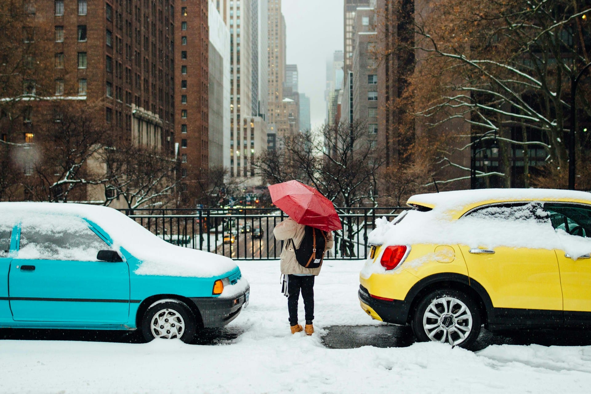 A blue and yellow car parked in the snow, a person with a red umbrella stands between them. 