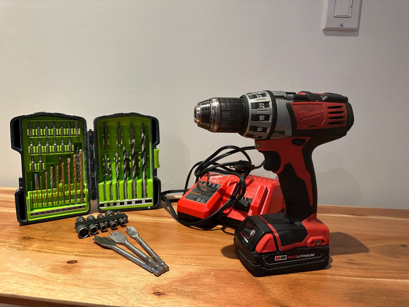 Perceuse-visseuse sans fil Milwaukee M18 avec chargeur, coffret d’embouts et mèches, forets plats et douilles, posés sur une table en bois