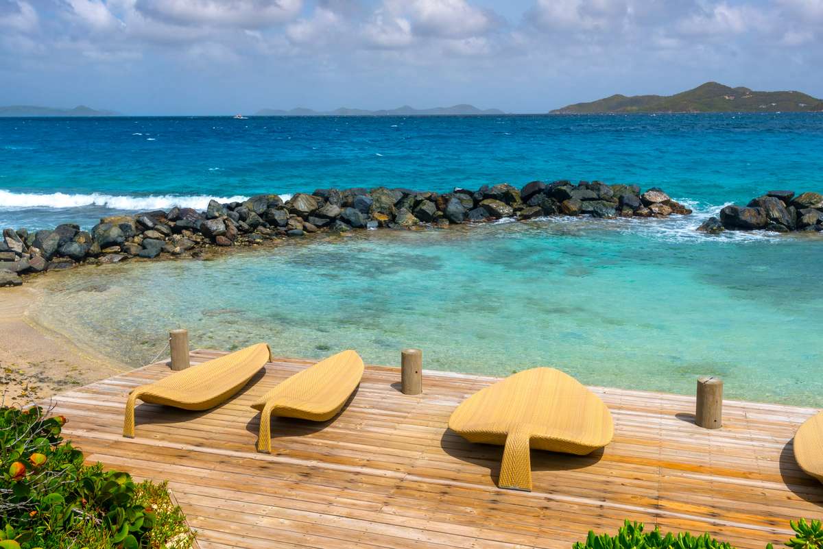 Three yellow lounge chairs sit on a wooden deck overlooking a turquoise lagoon with a rocky breakwater and ocean.