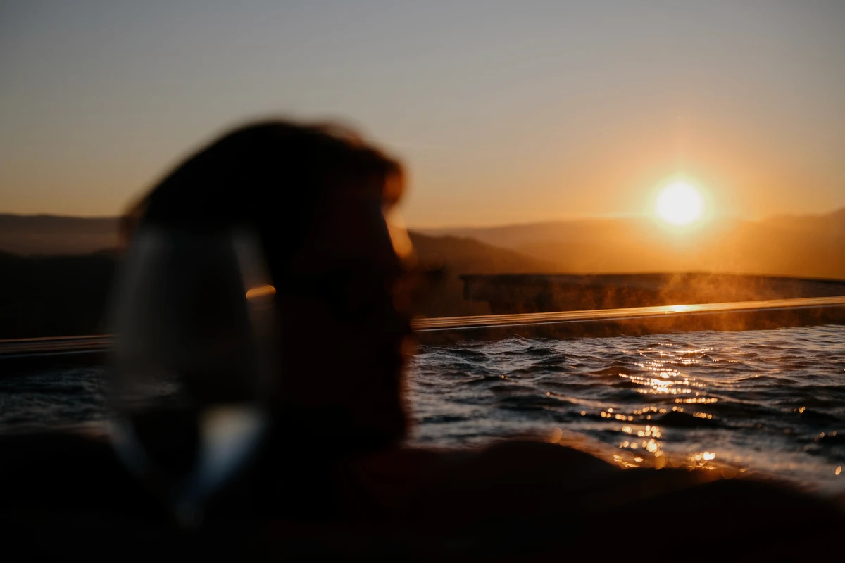 Gast blickt aus dem Pool in den Sonnenuntergang, im Vordergrund ein unscharfes Weinglas