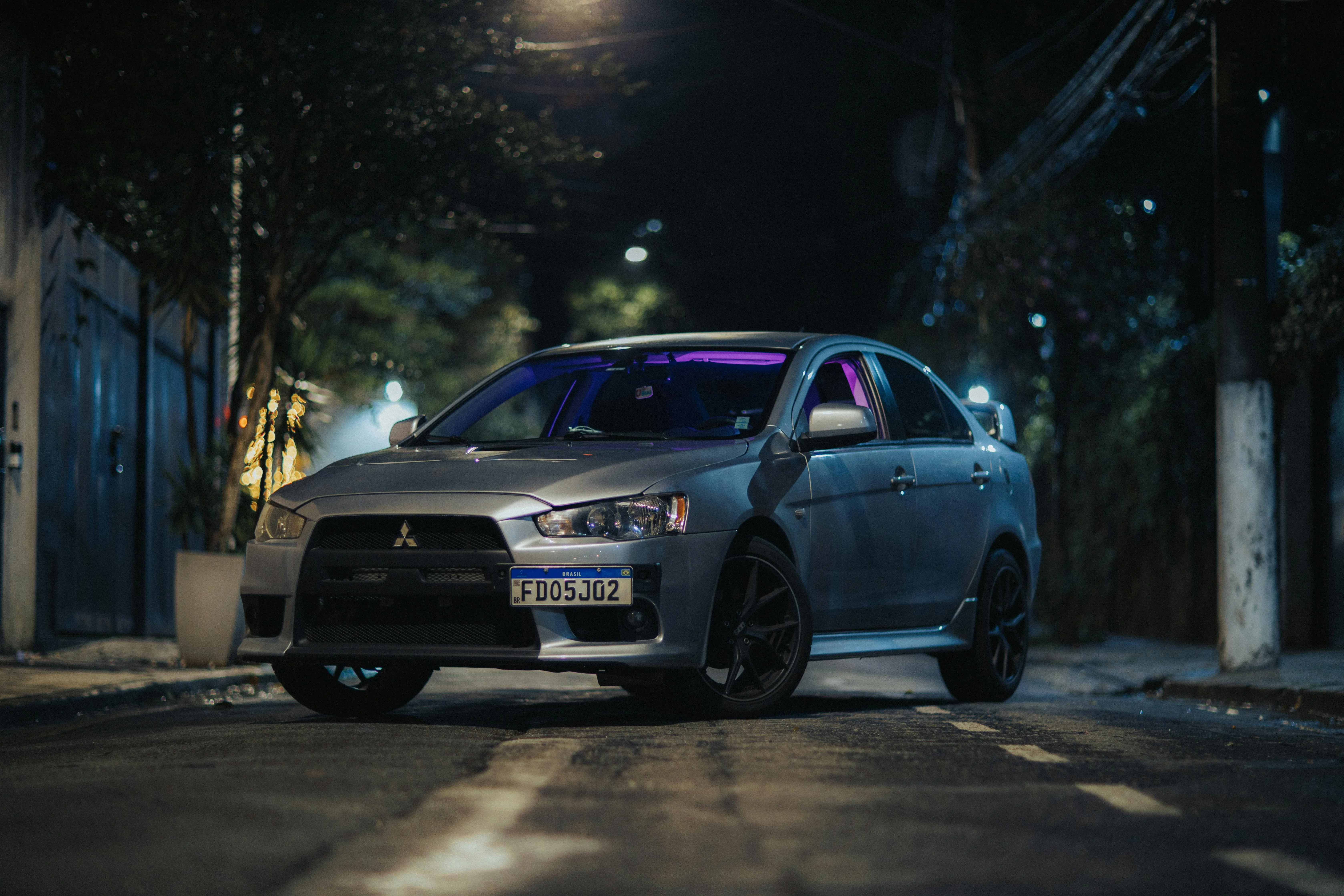 Une Mitsubishi Lancer photographiée de nuit dans une rue urbaine, éclairée par des reflets violets à l’intérieur. Sa posture basse, son bouclier avant massif et ses jantes sombres renforcent son caractère sportif, tandis que l’ambiance nocturne met en valeur son style agressif et précis.