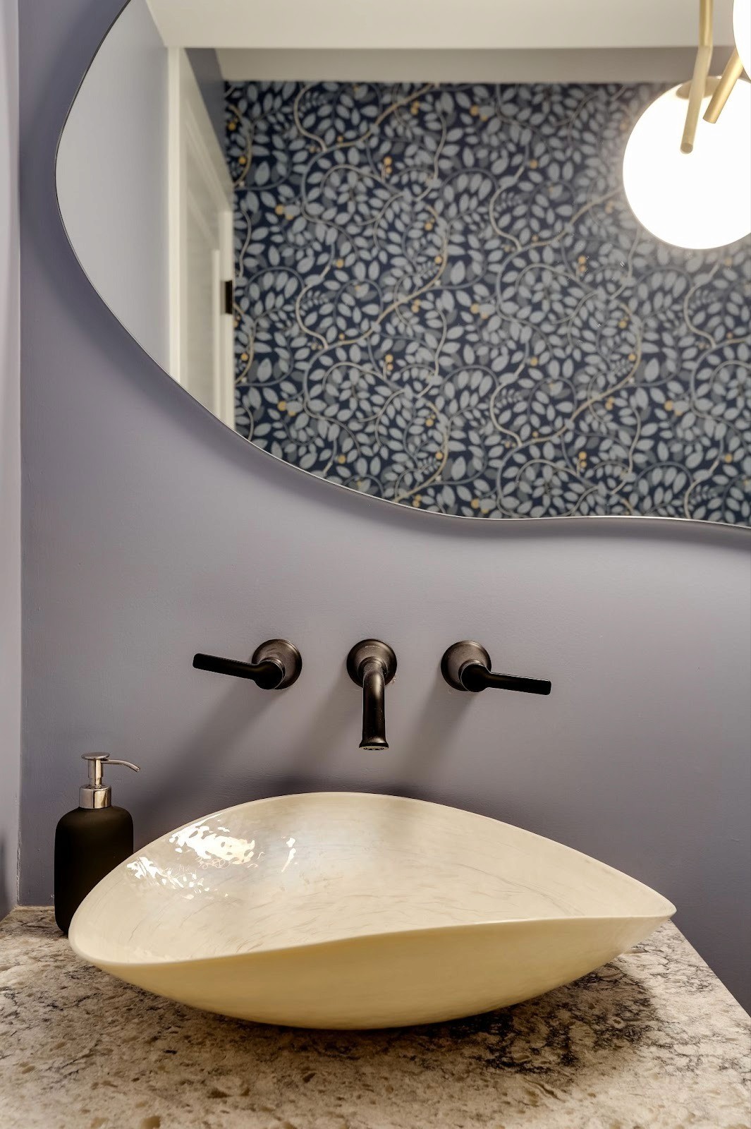 Close up of a modern powder room vanity with a sculptural vessel sink, wall mounted matte black faucet, curved mirror edge, and a blue patterned wallpaper reflected in the background.