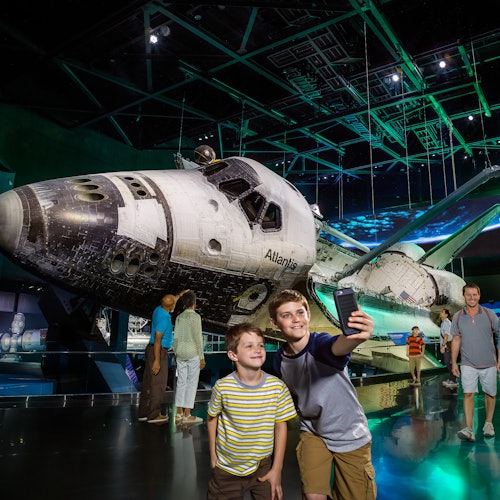 Två pojkar tar en selfie framför rymdfärjan Atlantis, som visas i ett museum med dramatisk belysning.
