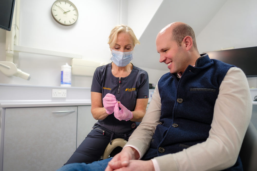 A female dentist named Beverley, wearing a face mask and pink gloves, holds a small dental implant and shows it to a bald male patient, who is sitting in a dental chair and smiling.