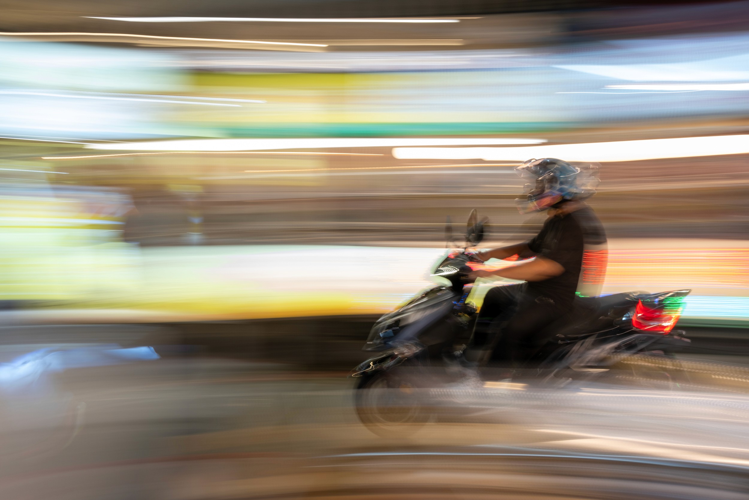 Color motion blur photograph of a person riding a scooter with bright streaks of storefront lights behind them.