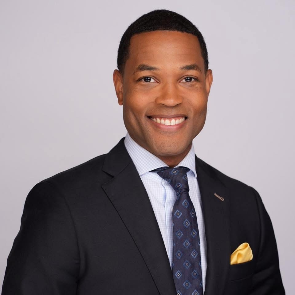 Mike Taylor in a dark suit, light blue dress shirt, and patterned tie smiles at the camera. He has short black hair and a yellow pocket square. The background is a plain light gray.