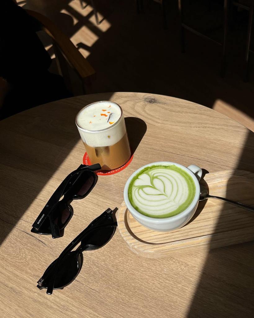 A top-down view of a wooden table featuring a glass of layered iced coffee with a foamy top and a matching green latte with intricate latte art, alongside a pair of stylish black sunglasses.