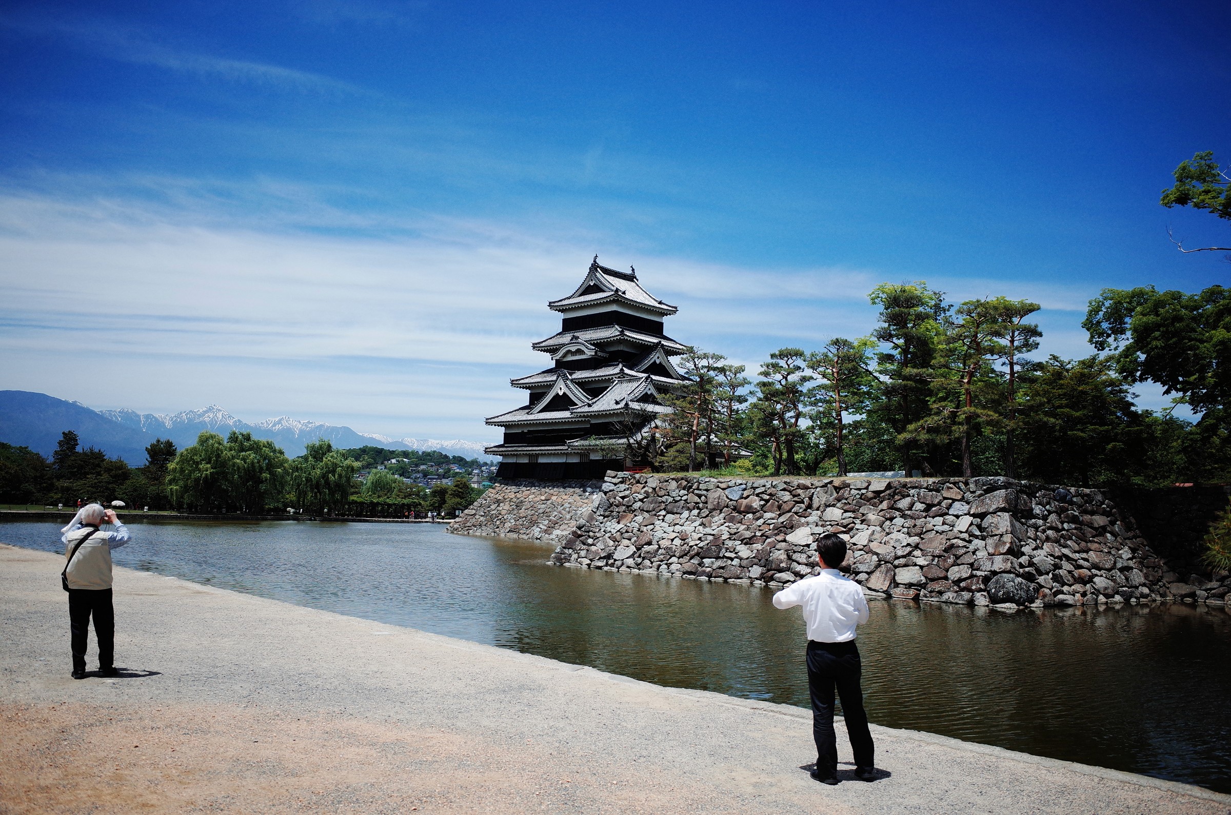 Photographie de voyage documentaire montrant 2 personnes japonaises se tenant au bord d'un lac photographiant un ancien chateau traditionnel japonais dans la ville de Matsumoto