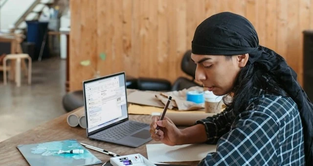 Pessoa de bandana escrevendo à mão ao lado de um laptop numa mesa de madeira, em escritório de pé-direito alto