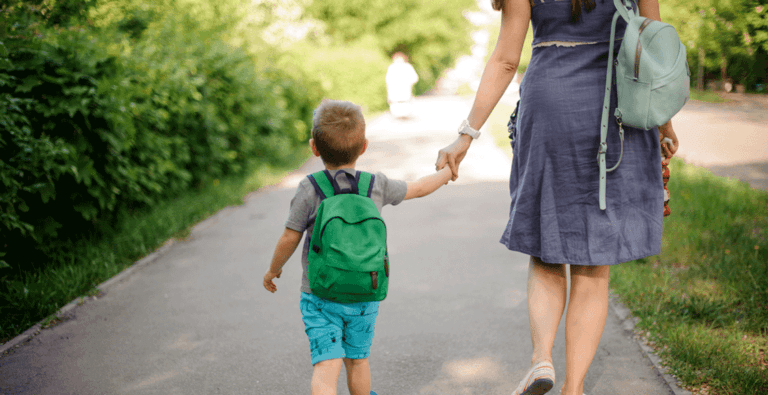 A child wearing a green backpack walking hand in hand with an adult along a green outdoor path
