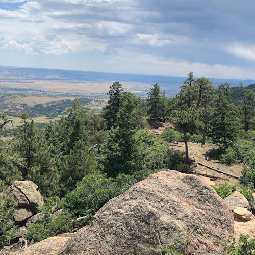 Mountain landscape with rocky foreground, dense green trees, and a distant plateau under a partly cloudy sky.