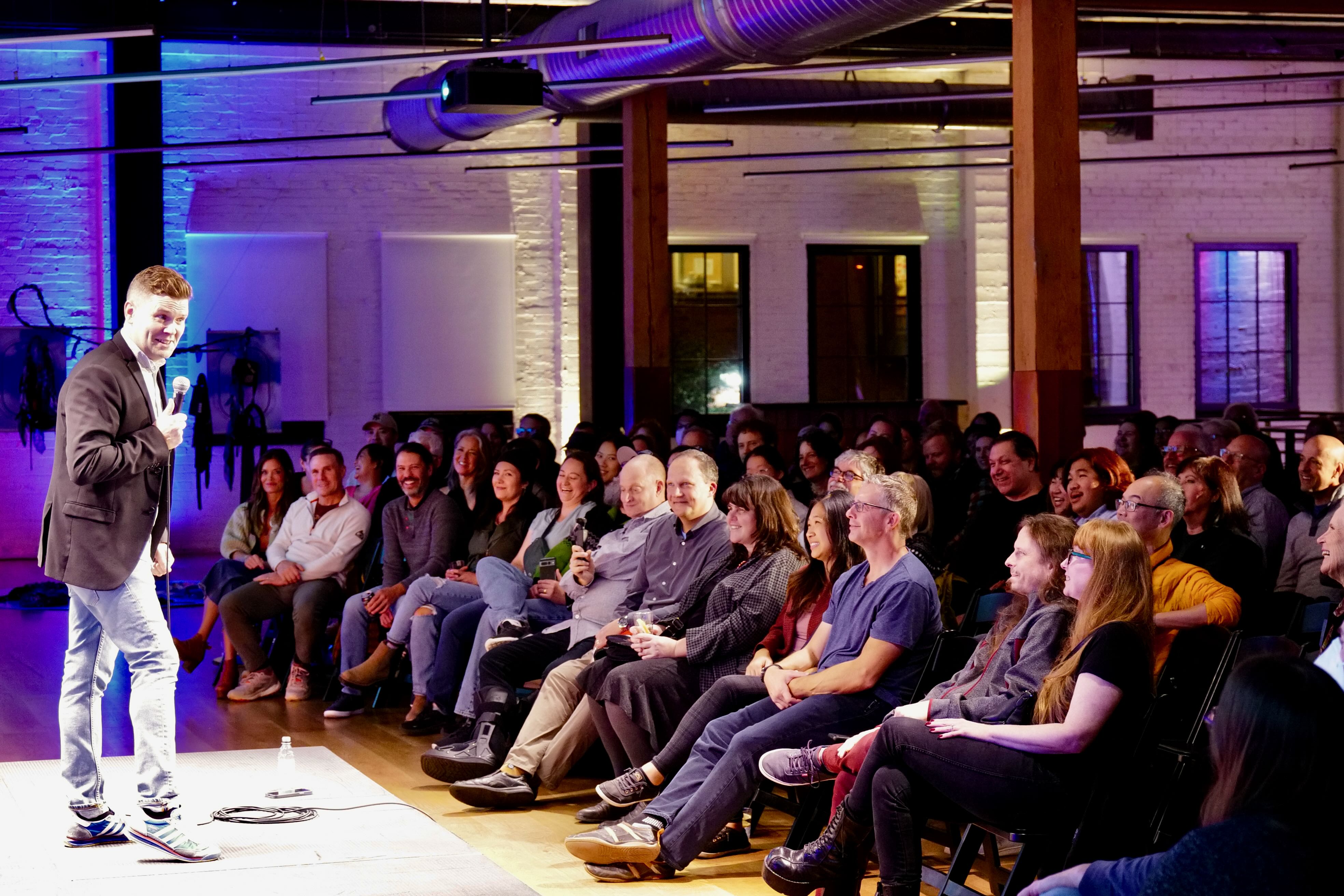 Icelandic comedian Ari Eldjárn stands on a brightly lit stage holding a microphone, performing a stand-up comedy set for a seated, smiling audience in an indoor venue with exposed brick walls and blue and purple accent lighting at Taste of Iceland Seattle.