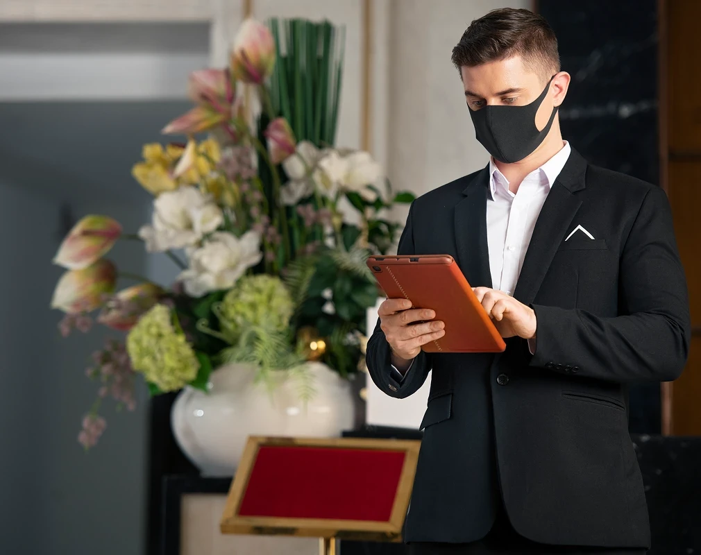 Мужчина в чёрном костюме с планшетом рядом с цветочной композицией в интерьере