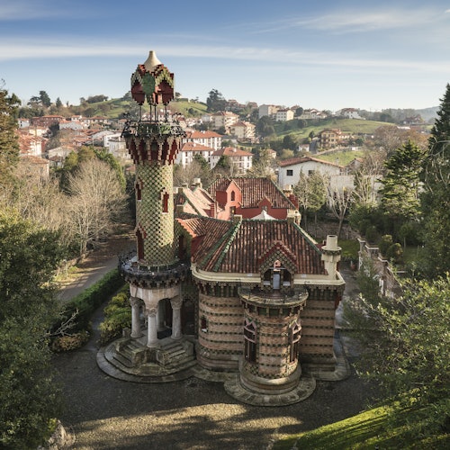 Un edificio caprichoso de varios pisos con techo de tejas y una torre decorativa, ubicado entre árboles con un pueblo y colinas al fondo.