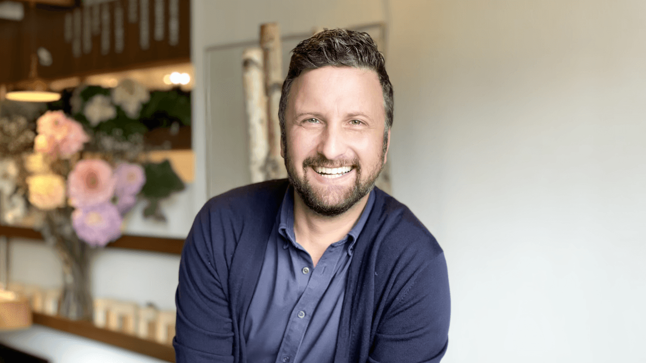A man with a cheerful expression and a well-groomed beard is the focus, wearing a navy blue cardigan over a matching shirt. He is smiling broadly, conveying warmth and friendliness. The background is softly blurred, featuring light-colored wooden branches upright in a minimalist setting. Soft natural lighting highlights his features, creating a warm and welcoming atmosphere. The image is composed with a close-up perspective, centered on the man's face, emphasizing his joyful demeanor.