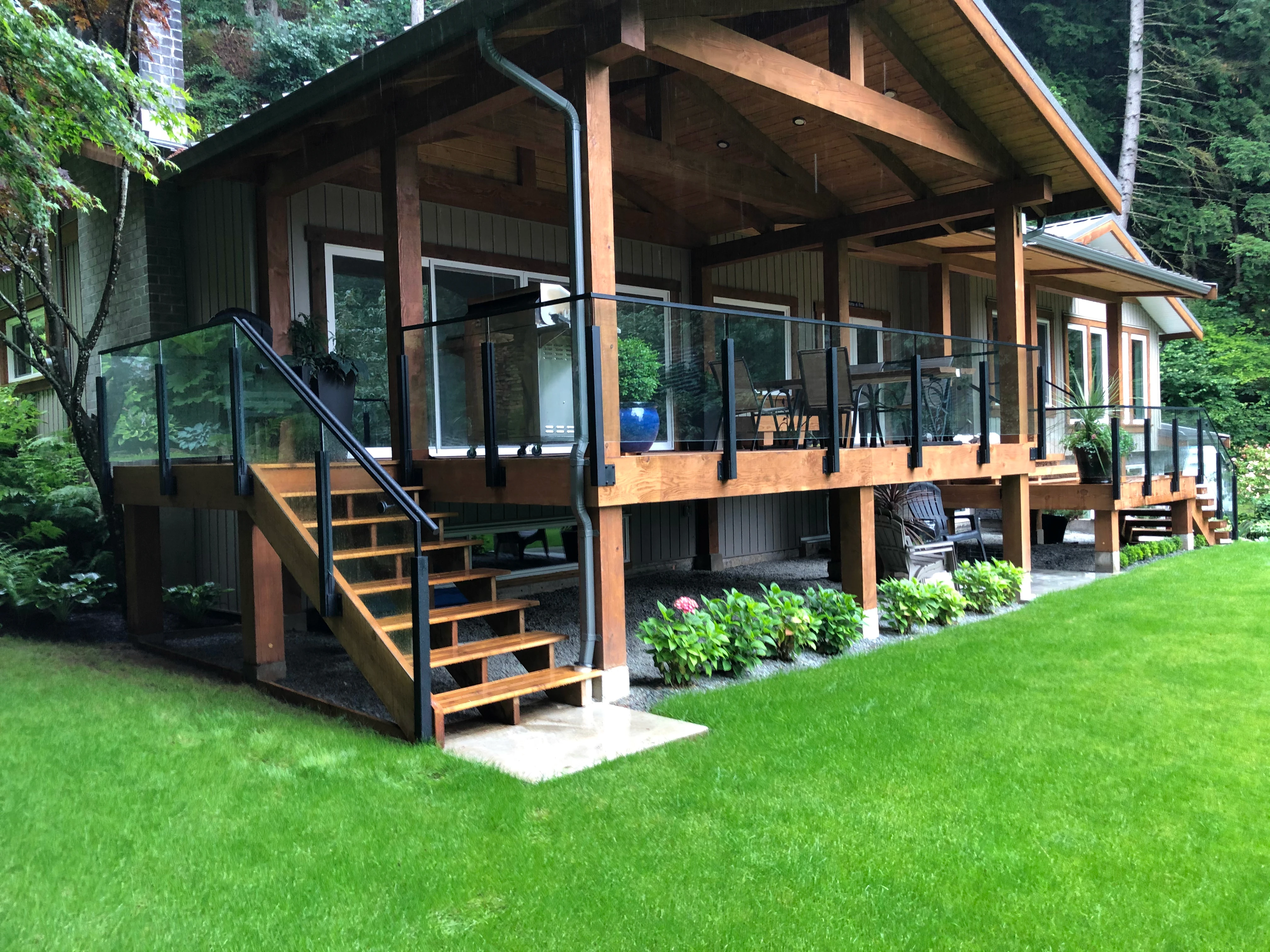 Modern timber-frame coastal home with frameless glass railings on an elevated cedar deck, surrounded by lush Pacific Northwest forest.