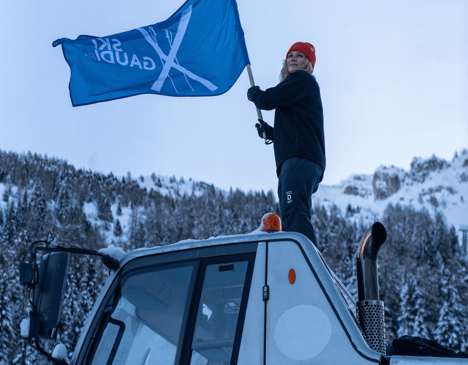 Person waving blue flag on snowcat in snowy mountain landscape.