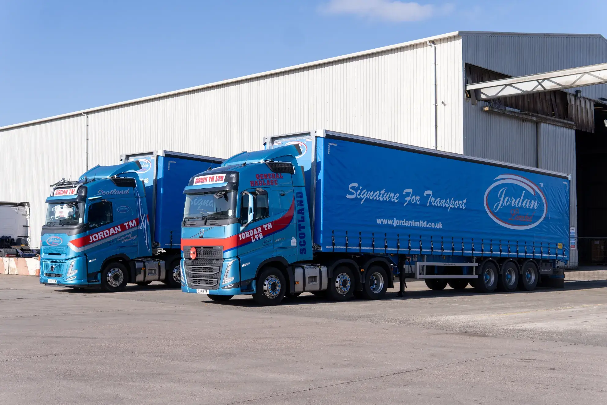 Two blue trucks with "Jordan TM" branding are parked outside a large warehouse on a sunny day. The trailers display "Signature for Transport."