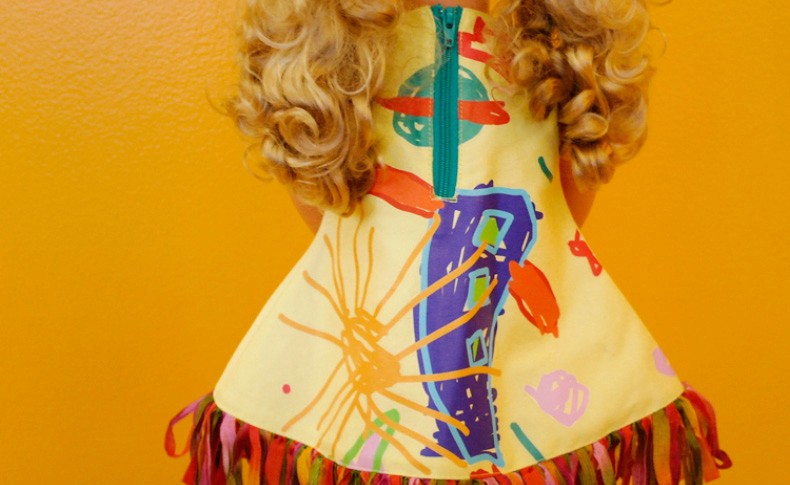 A person with curly blonde hair stands against an orange wall, wearing a yellow sleeveless top. The top features a colorful abstract design including a blue shape with radiating lines and smaller shapes in red, orange, and pink. The bottom hem of the top has multicolored fringed fabric. The person’s back is to the camera, and their head is turned slightly to the right.