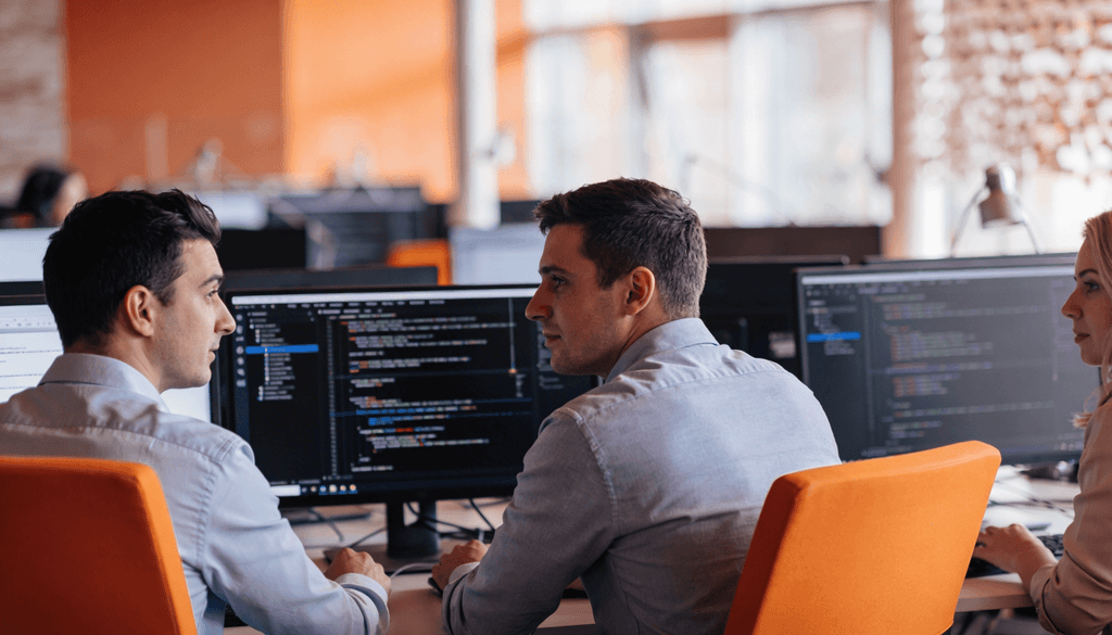 Equipe de tecnologia colaborando em frente a monitores com código, em escritório moderno com iluminação laranja.