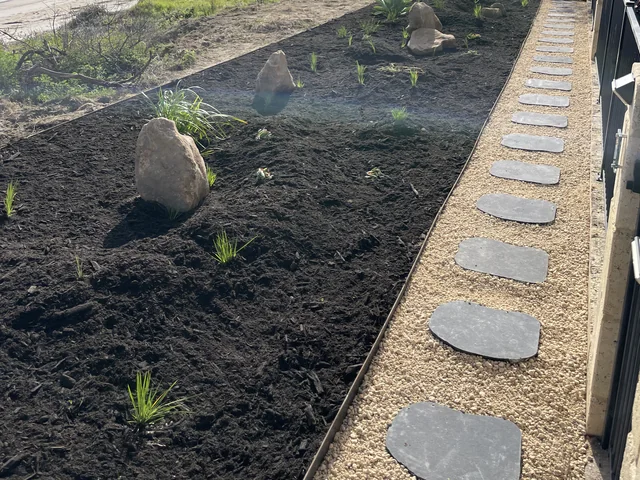 Dark stepping stones in pale gravel beside a newly planted, steel-edged rock garden.