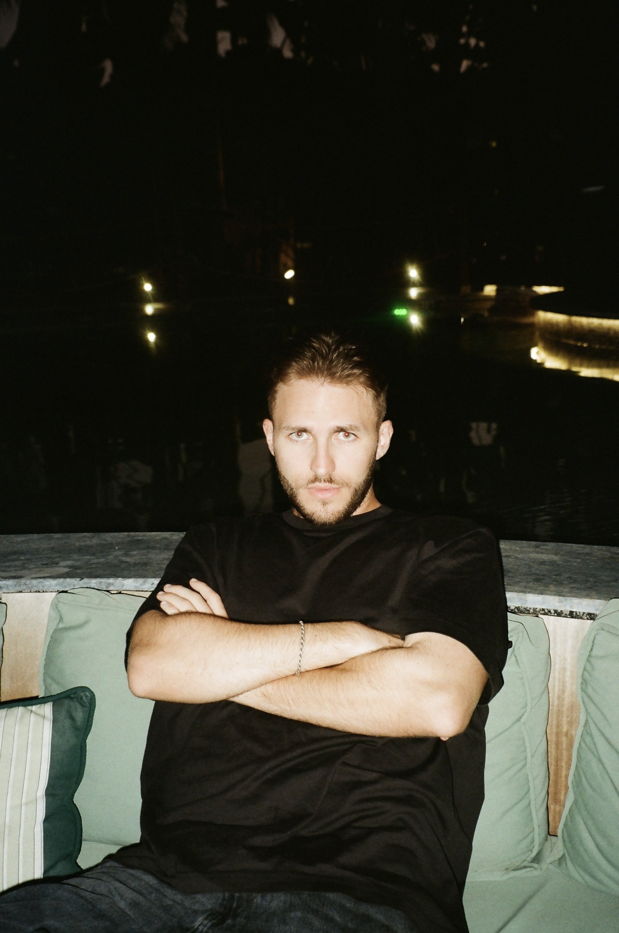 A portrait of Polish music producer Hubi sitting outdoors at night. He is wearing a black t-shirt, with his arms crossed and a confident expression, against a dark, illuminated background.