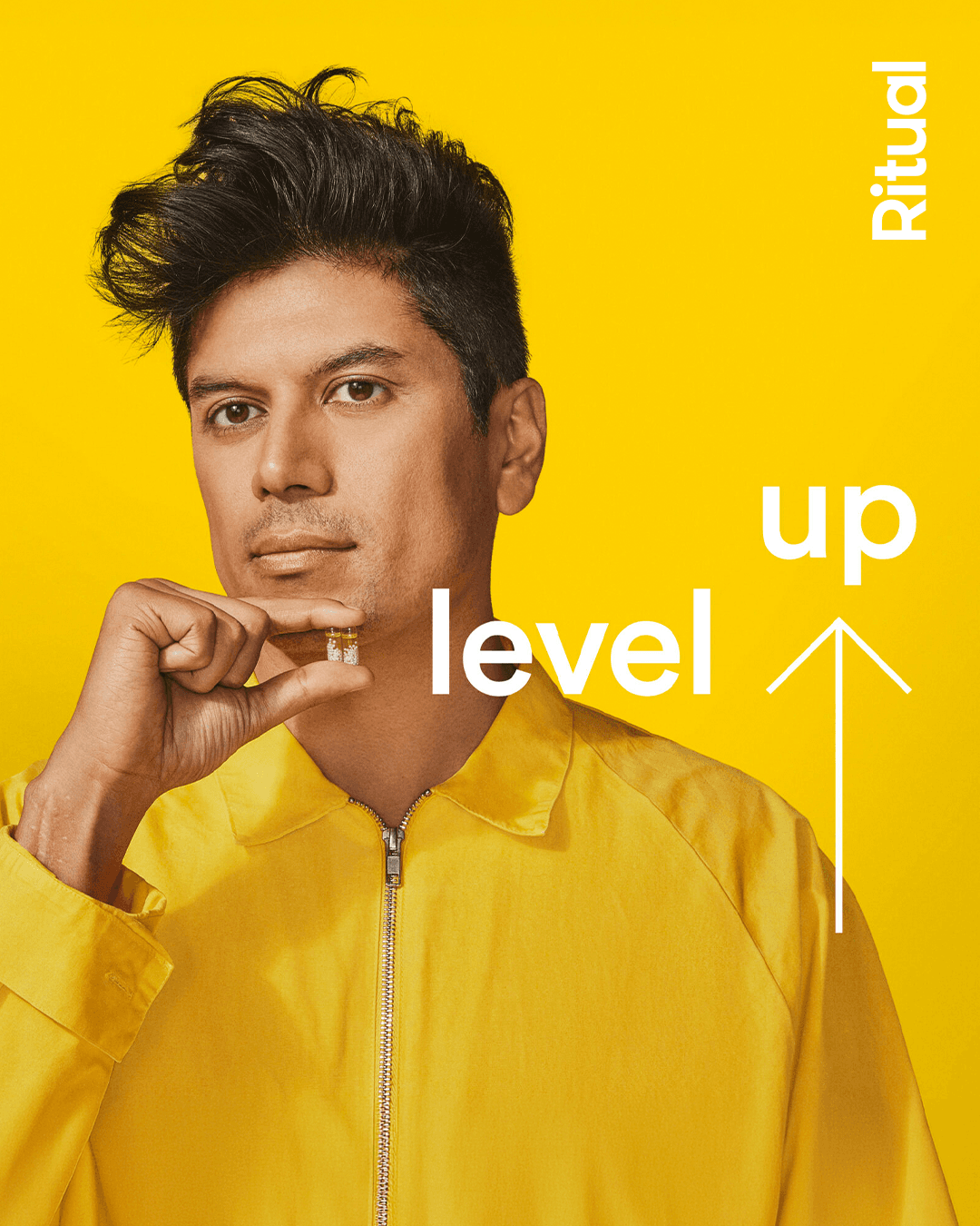 Ritual branded image of a man holding two Ritual vitamin capsules. Wearing a yellow jacket, in front of a yellow background, this image exudes Ritual brand.
