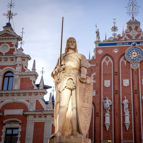 一座骑士雕像持剑盾，伫立在装饰华丽、历史悠久且细节复杂的建筑前，伴随着一个时钟。