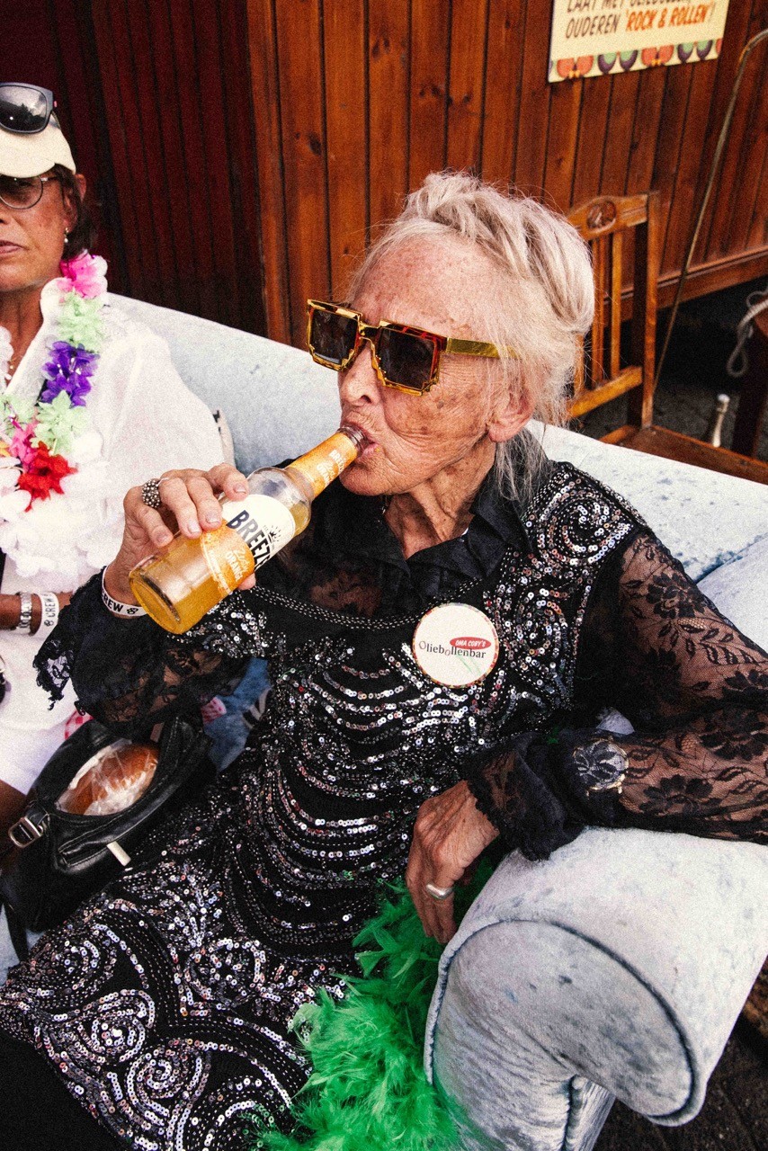 Oudere vrouw met glitterjurk en zonnebril drinkt een Breezer op een feestje