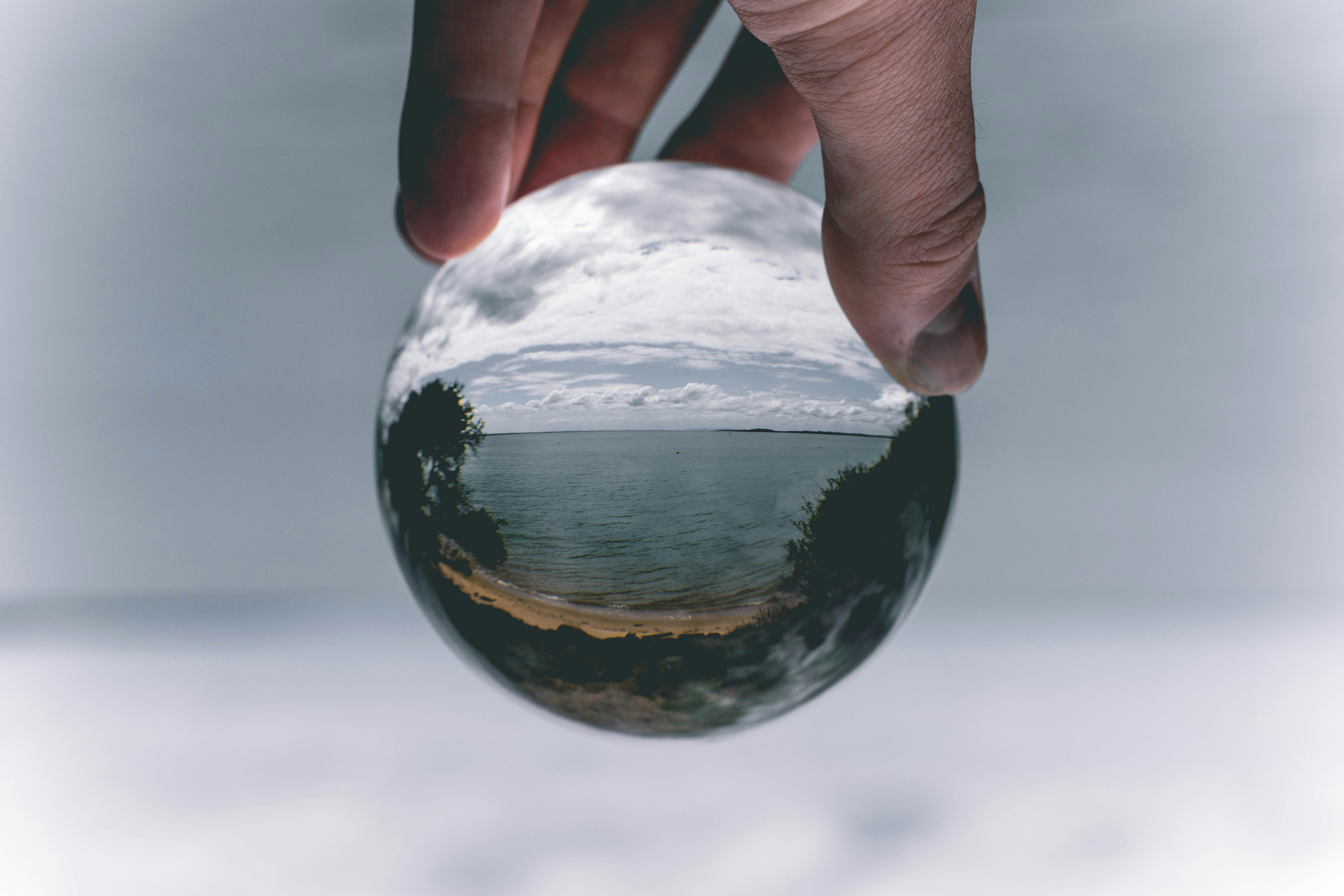 A hand holds a glass orb, reflecting a scenic view of water and clouds in its surface.