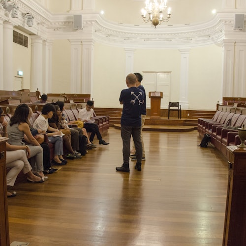 People seated in a large, ornate room with wooden floors and white walls, two individuals standing and talking near the front.