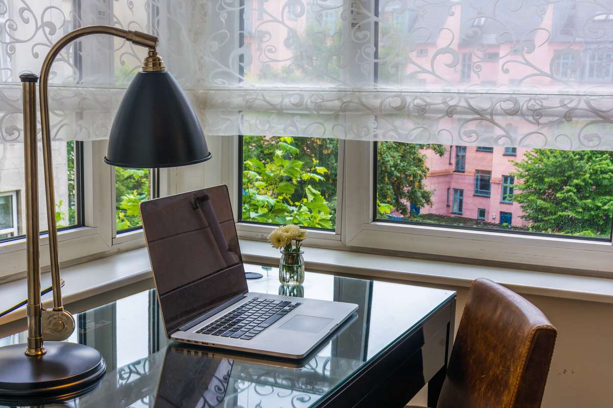 A laptop and a small vase of flowers sit on a dark desk in front of a large window with white lace curtains.