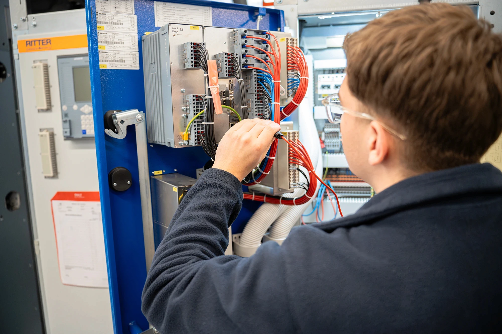 Ein Mitarbeiter von RITTER Starkstromtechnik prüft die Verdrahtung und elektrische Komponenten in einem geöffneten Schaltschrank. Er führt eine Kontrolle der Elektroanlage im Produktionsbereich durch.