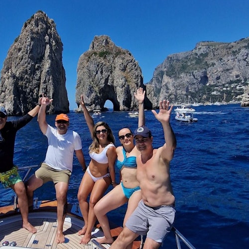 Five people on a boat, raising their hands, with rocky cliffs and boats in the background under a clear blue sky.