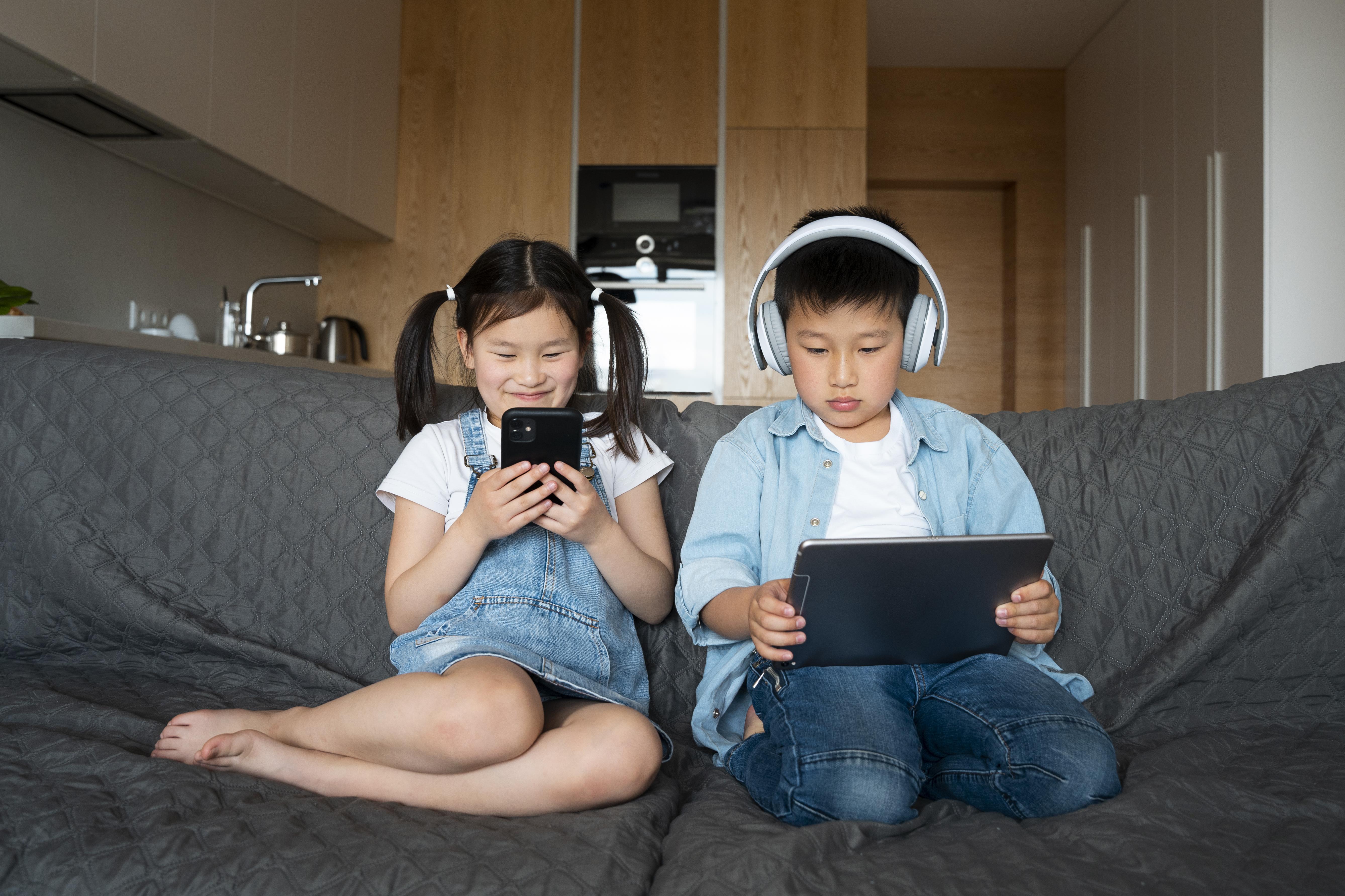 Two children sitting on a couch; a girl holding a smartphone smiles, while a boy wearing headphones focuses on a tablet. Cozy home setting.