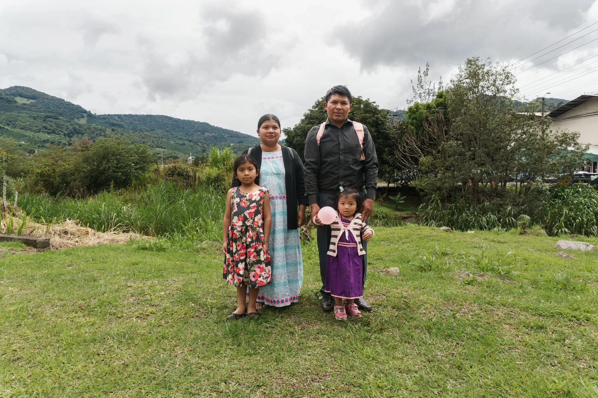 A family of four stands on a grassy field with mountains in the background. The father holds a backpack, the mother wears a long dress, and the two young girls stand in front, wearing colorful dresses, holding pink balloons.