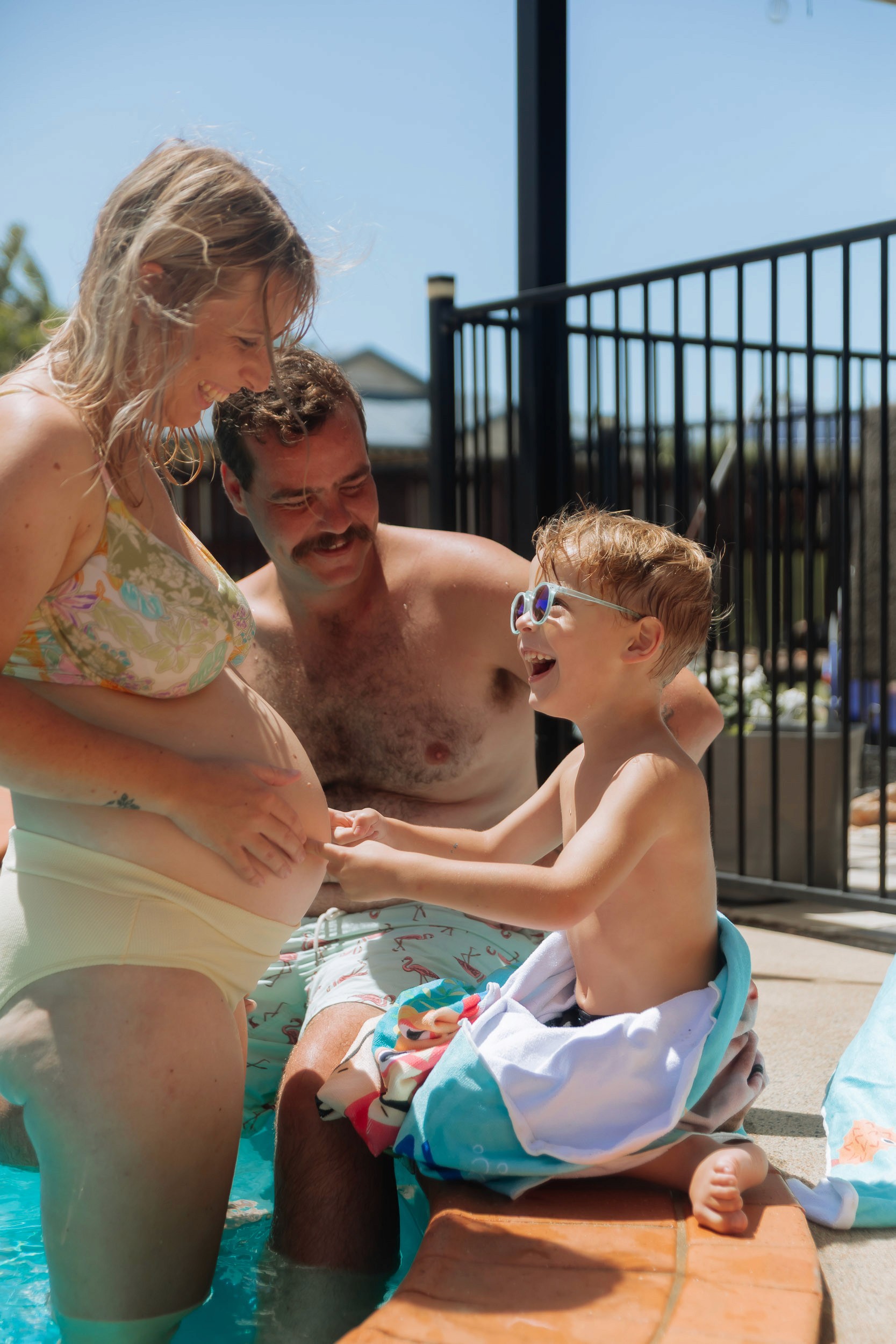 Candid poolside family moment with an expectant mother, dad, and child laughing together in the backyard pool.
