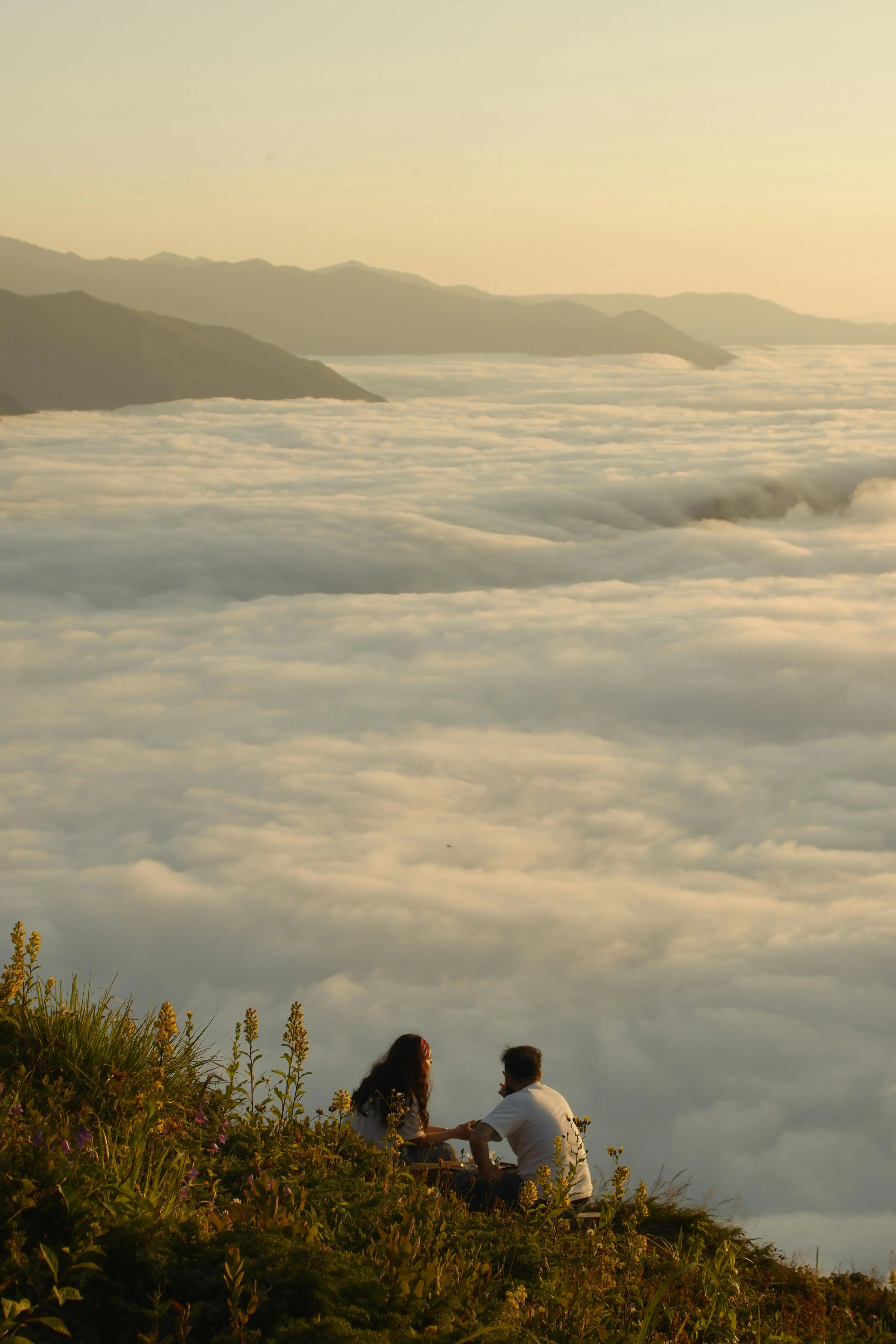 Two people sit on grassy ground overlooking a sea of clouds with mountains in the background during golden hour.