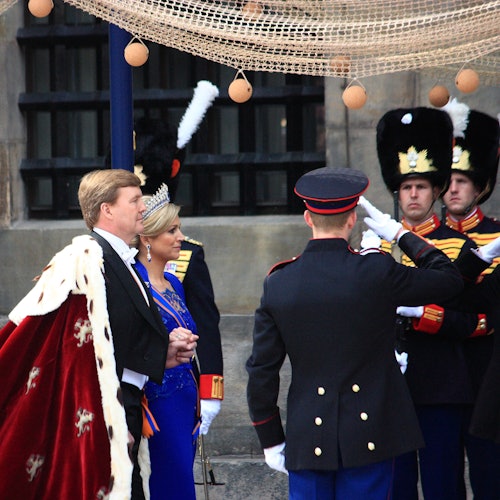 En man i en röd och vit ceremoniell dräkt och en kvinna i en blå klänning med tiara står tillsammans med soldater i utarbetade uniformer.