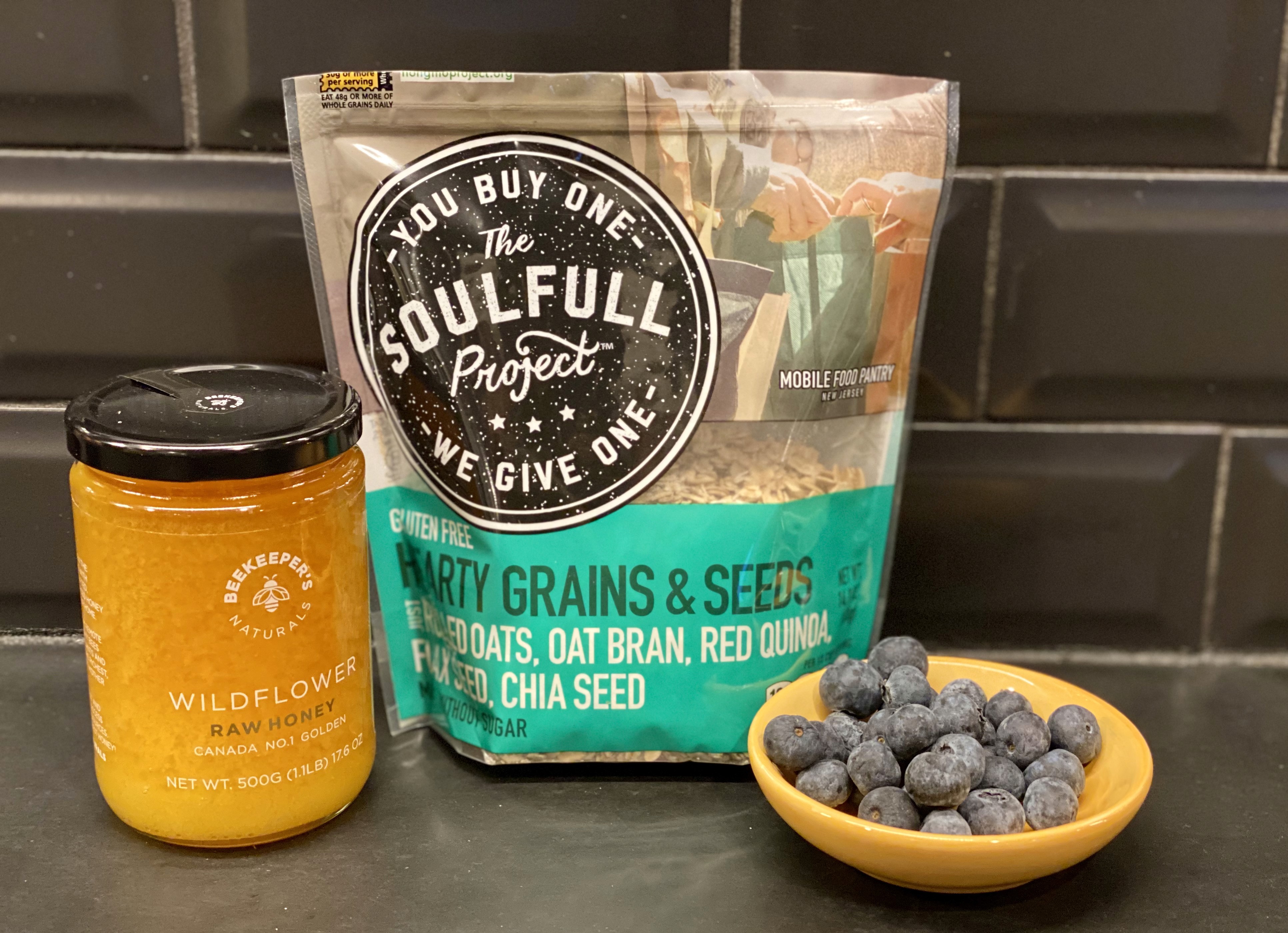 A jar of Beekeeper's Naturals Wildflower Raw Honey stands beside a bag of The Soulful Project Hearty Grains & Seeds, with fresh blueberries scattered on a dark countertop, against a black tiled backsplash.