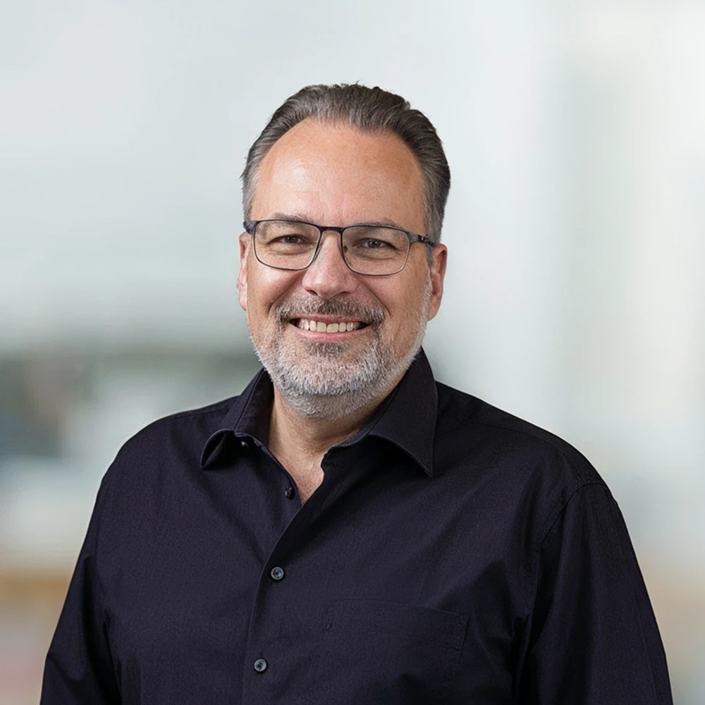 A smiling man with glasses wearing a black shirt, standing in a well-lit, blurred background.