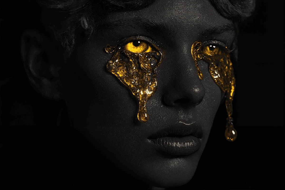 A close-up of a person's face with shimmering liquid gold dripping from their eyes and across their skin.