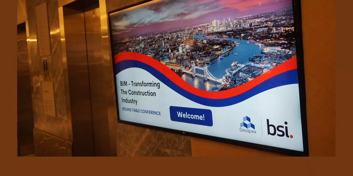 A man stands while two people sit at a table in an office setting, smiling and engaged in conversation. The mood is friendly and collaborative.