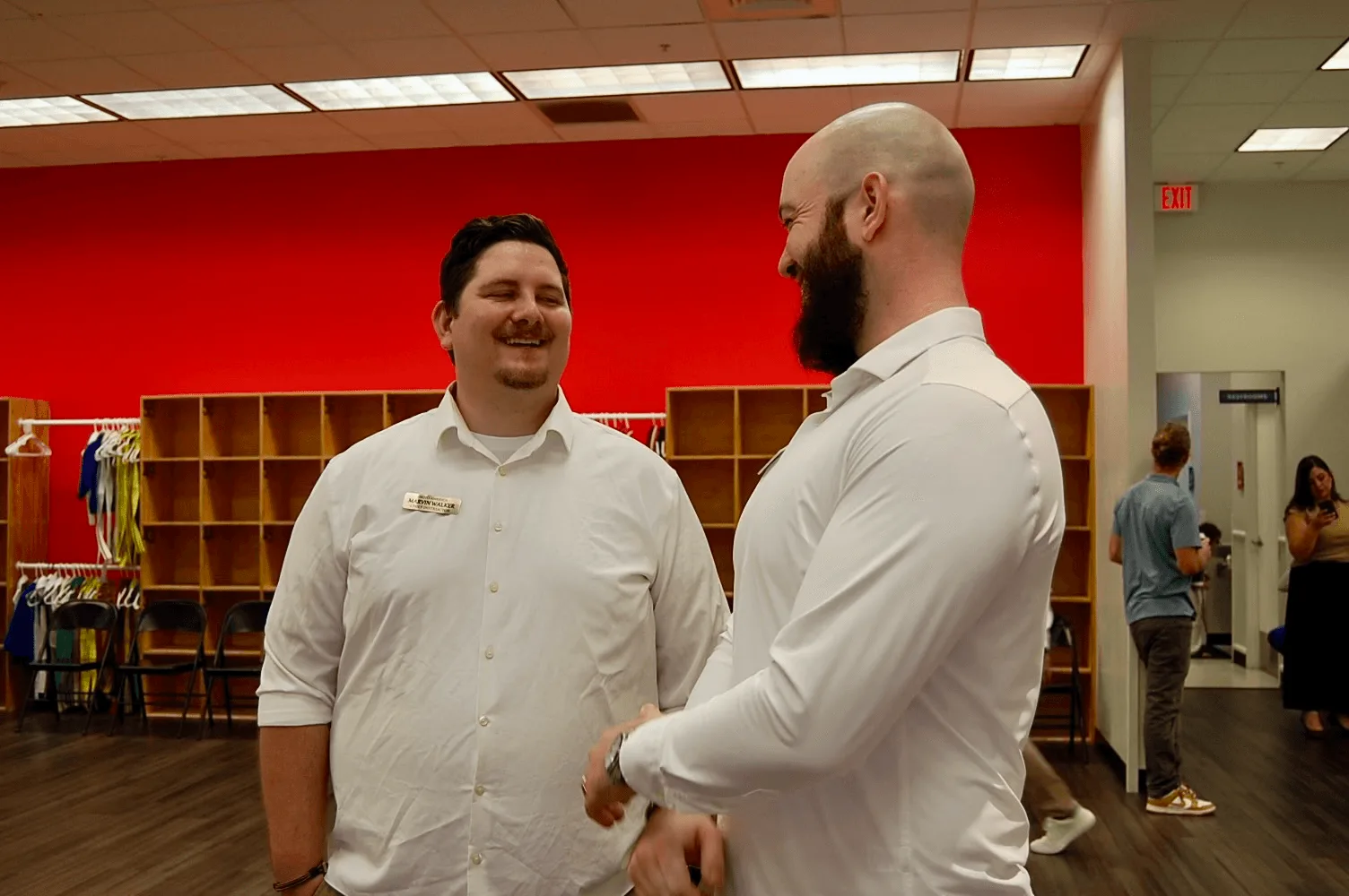 Two men in white shirts shake hands and smile while conversing in a library or community center with bright red walls, wooden shelving, and other people visible in the background.